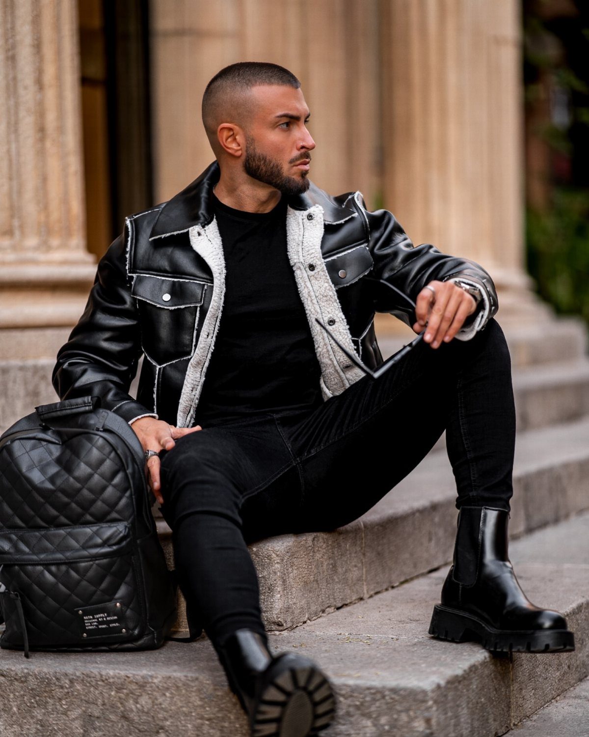 Man in a black leather shearling coat and chunky boots sitting on stone steps.