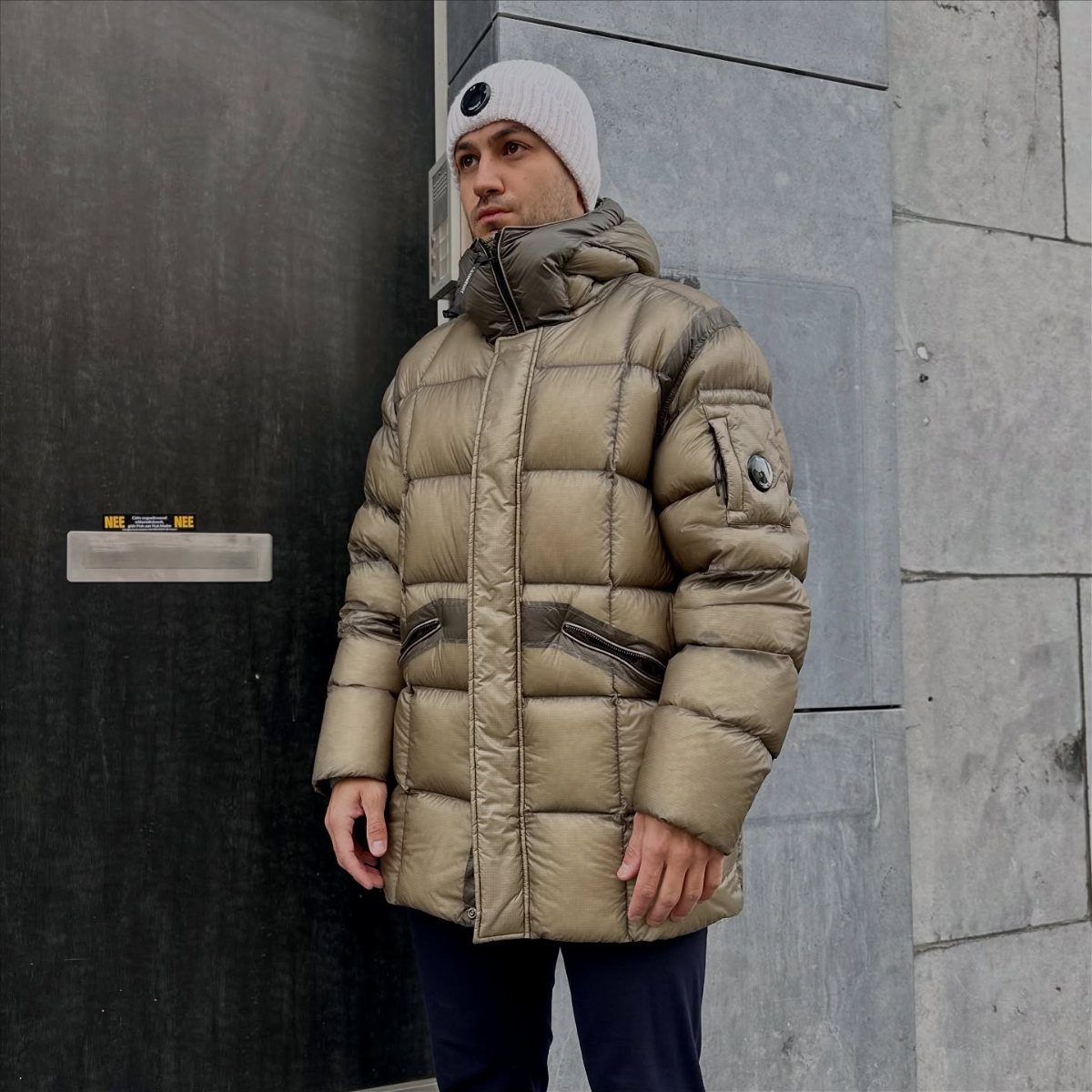 Man in a tan puffer coat and white beanie standing by a stone wall.