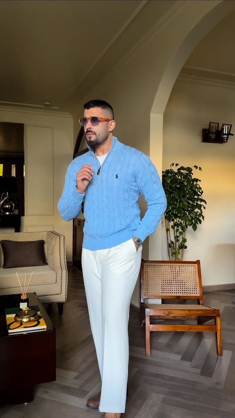 Man wearing a light blue cable-knit sweater and white trousers in a living room.