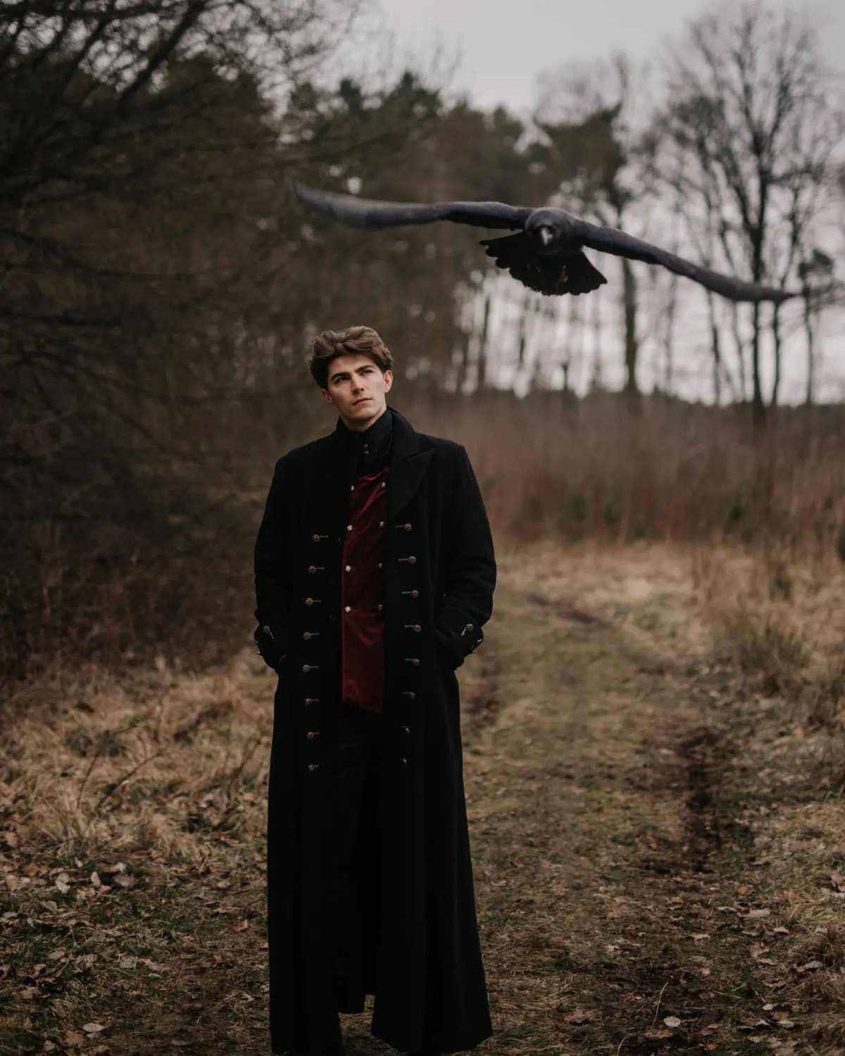 Man in a long black coat and red vest standing in a winter field.