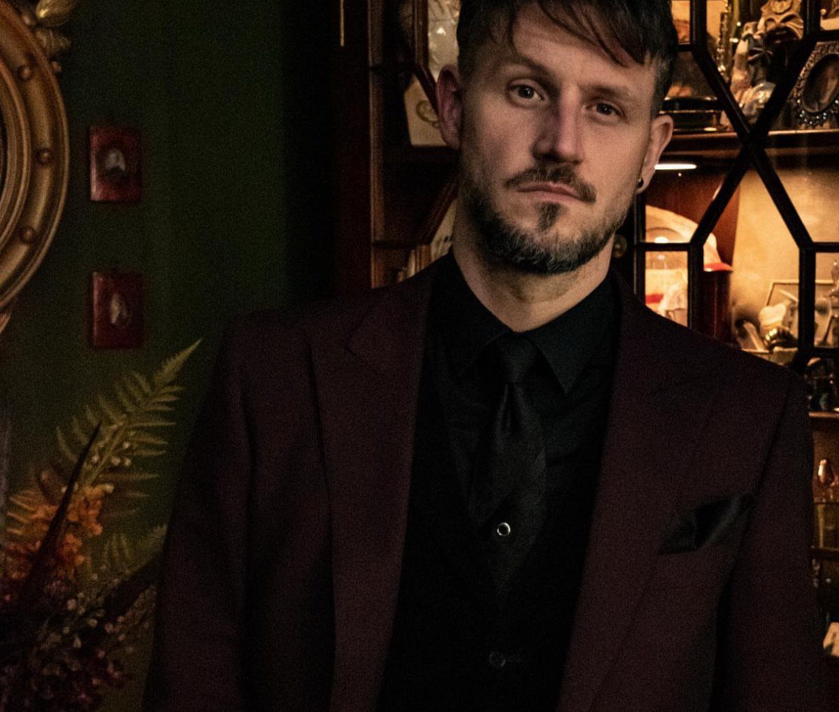 Man wearing a burgundy blazer and black shirt in a dark, gothic setting.
