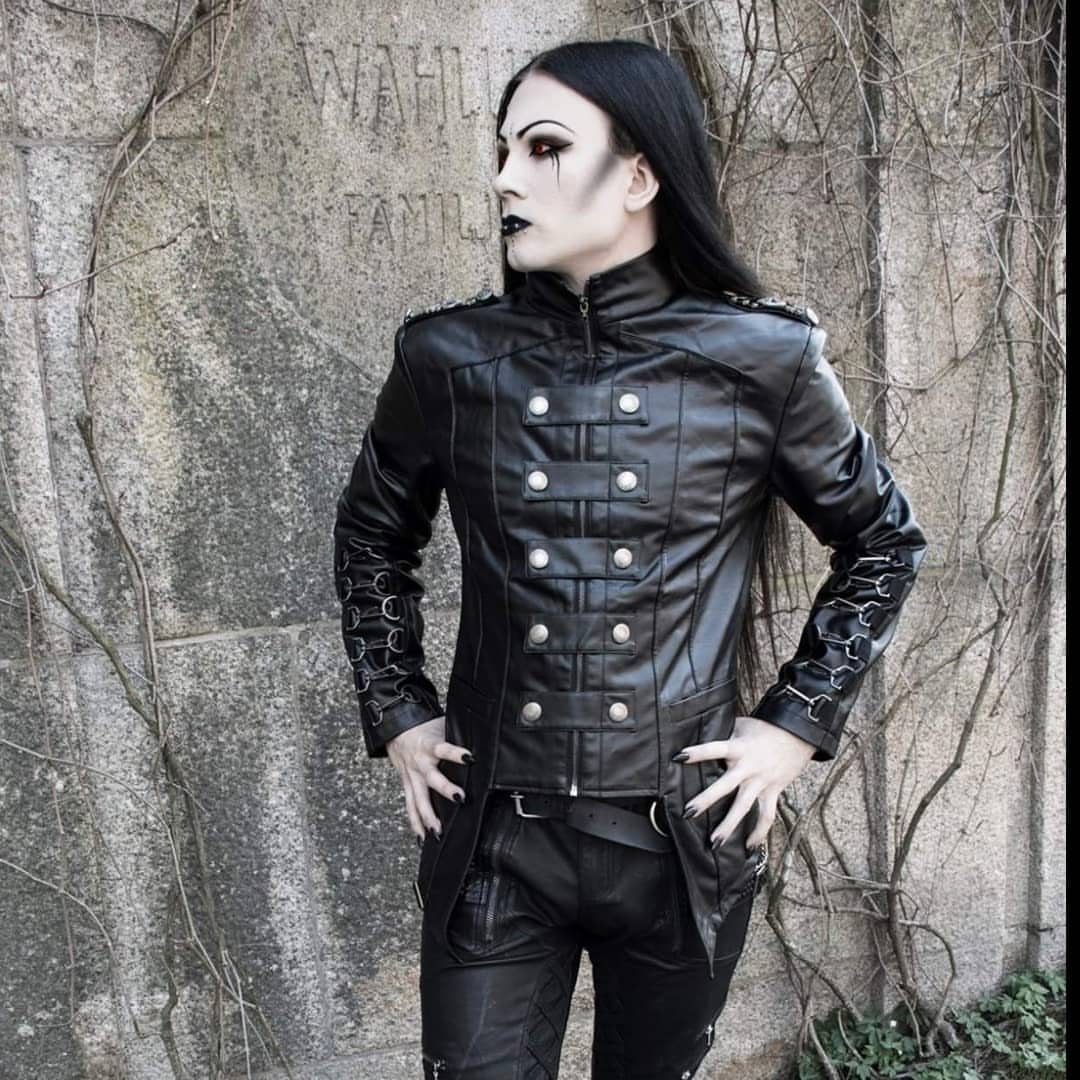 Man in black leather military jacket and gothic makeup posing by a stone wall.