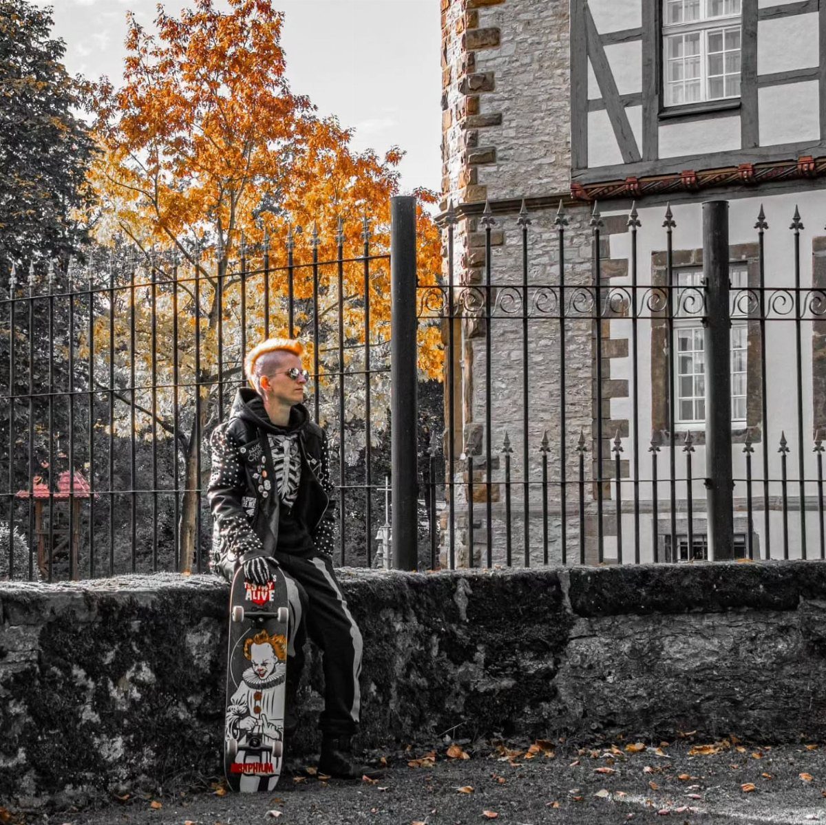 Man with orange hair in studded leather jacket and black clothing holding a skateboard.