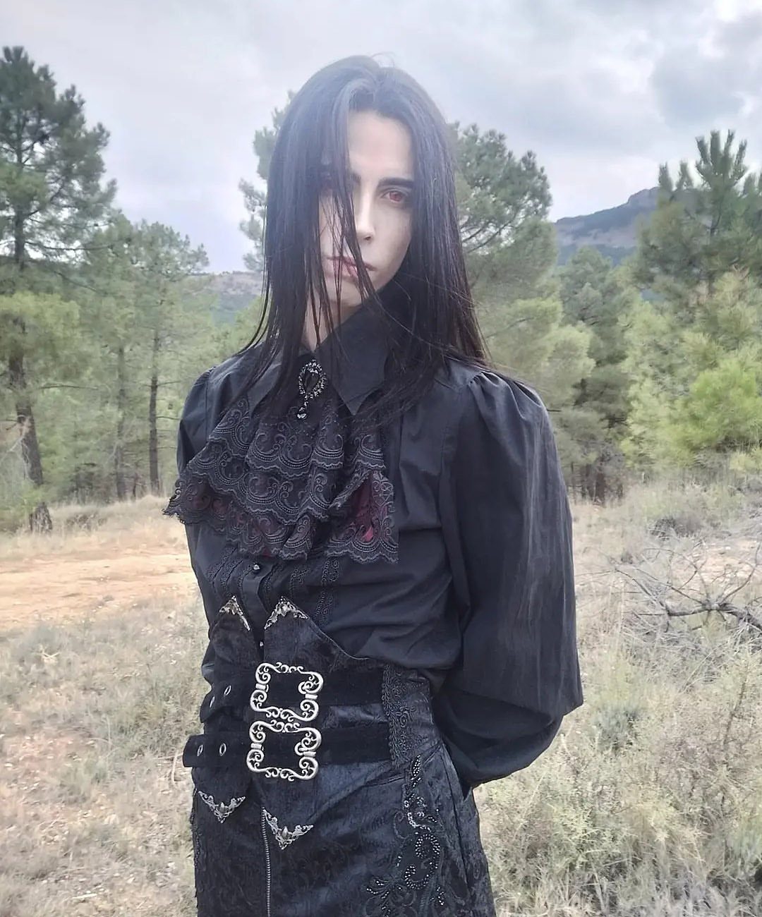 Person in gothic black attire with lace ruffles and ornate belt in a forest