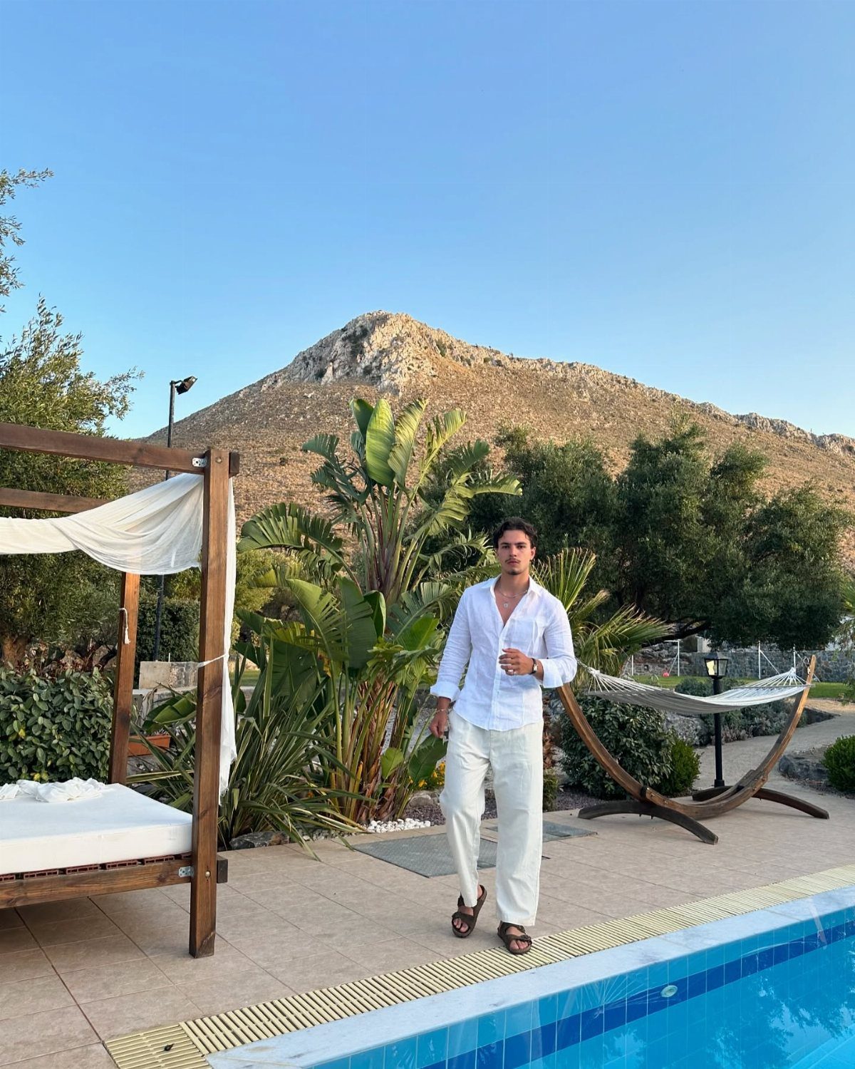 Man in white linen shirt and trousers standing by a pool with mountains behind.