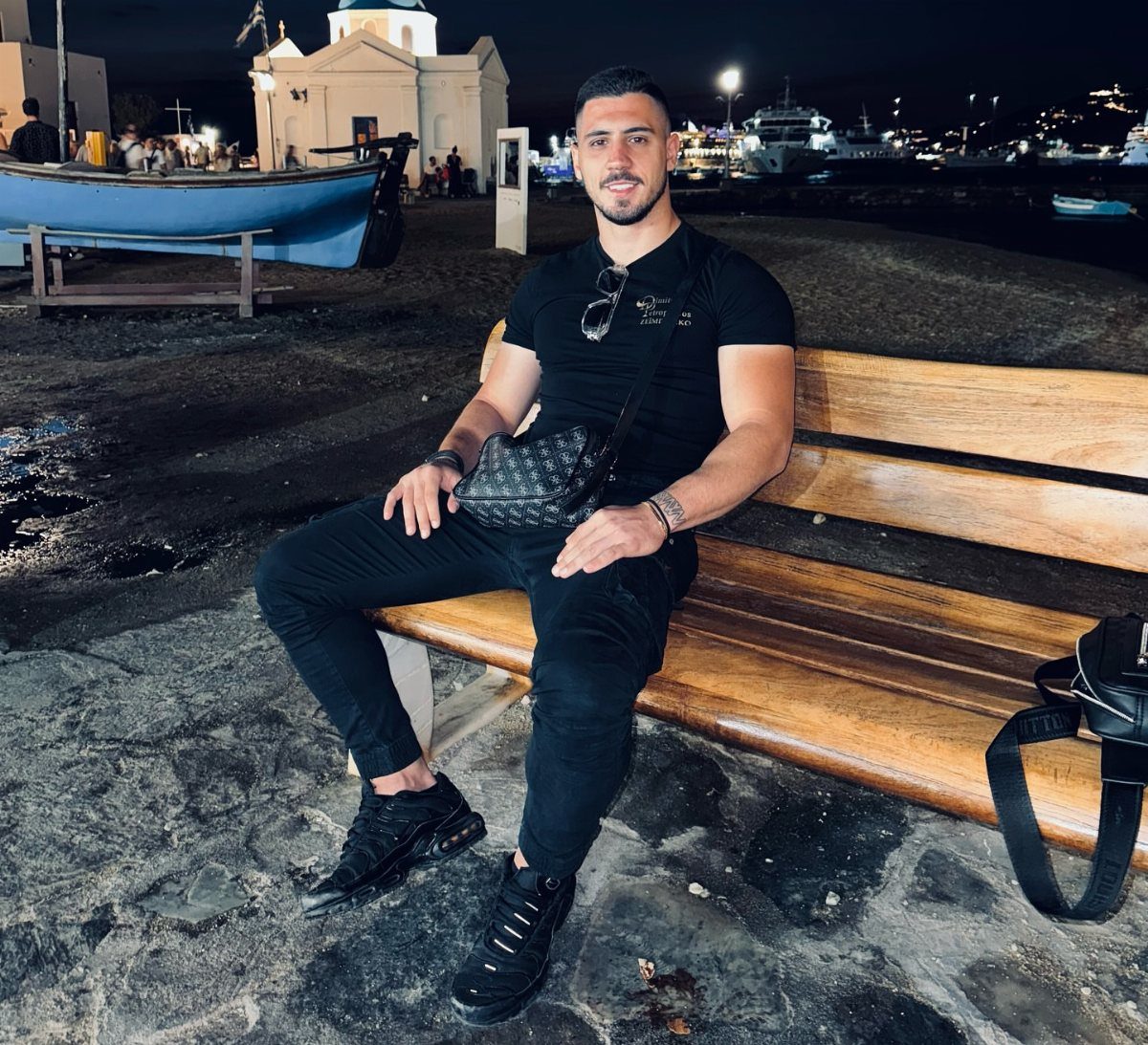 Man in all-black outfit sitting on a wooden bench by a harbor at night