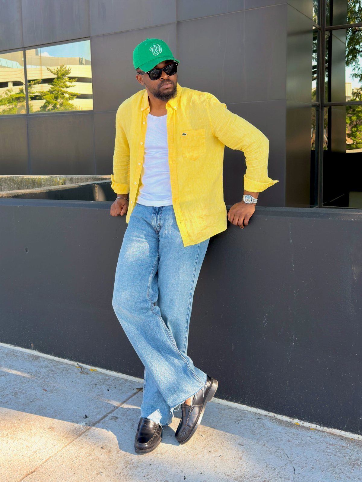 Man in yellow shirt, light blue jeans, and green cap leaning against wall.