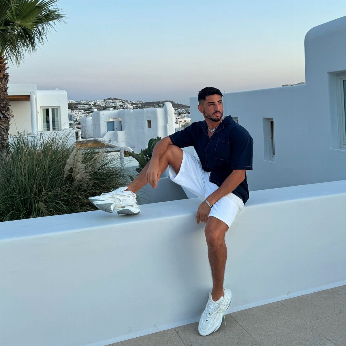 Man wearing a navy blue shirt and white shorts sitting on a white wall.
