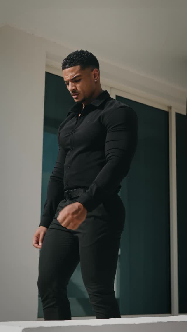 Muscular man wearing a fitted black button-down shirt and black trousers posing indoors.