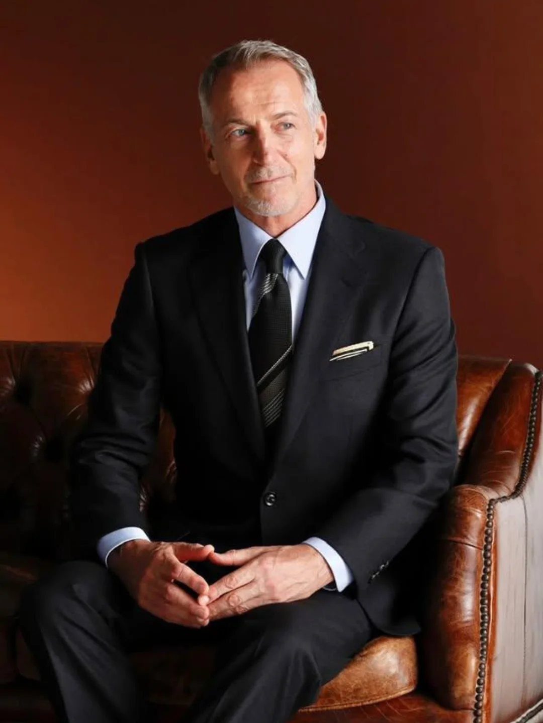 Man in a black tailored suit and tie sitting on a leather sofa.