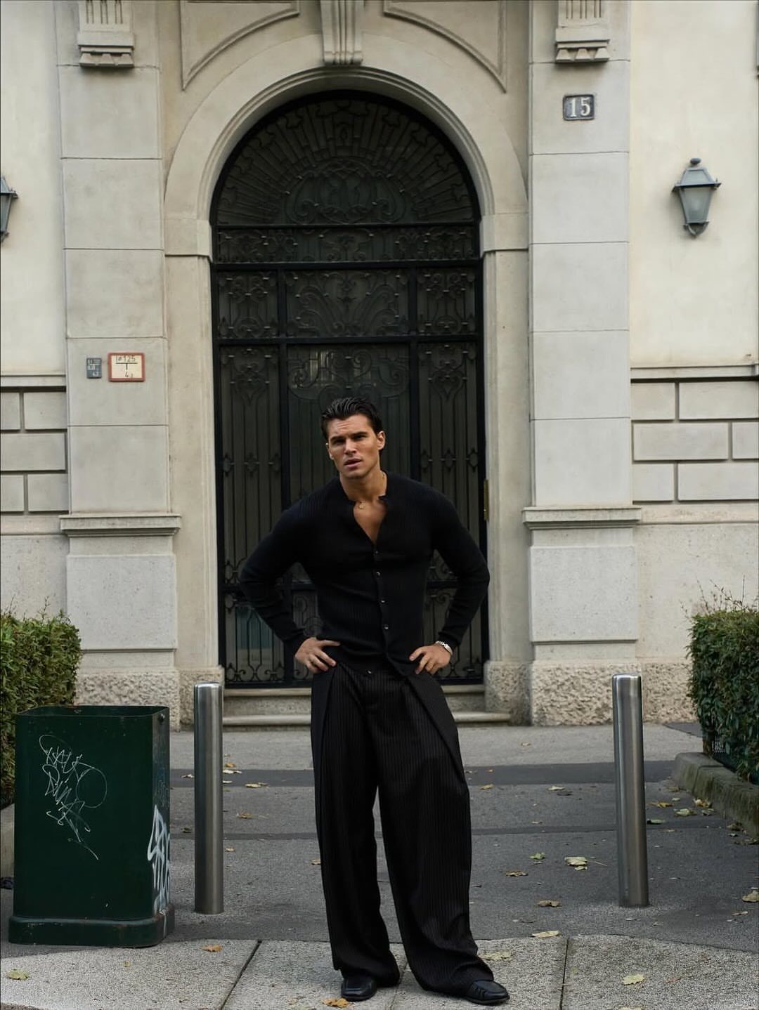Man in tight black shirt and wide black trousers posing by arched door