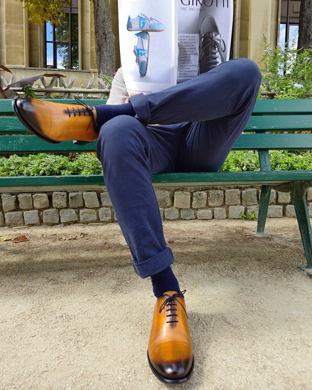 Polished tan leather oxford shoes worn with navy blue trousers on a park bench.