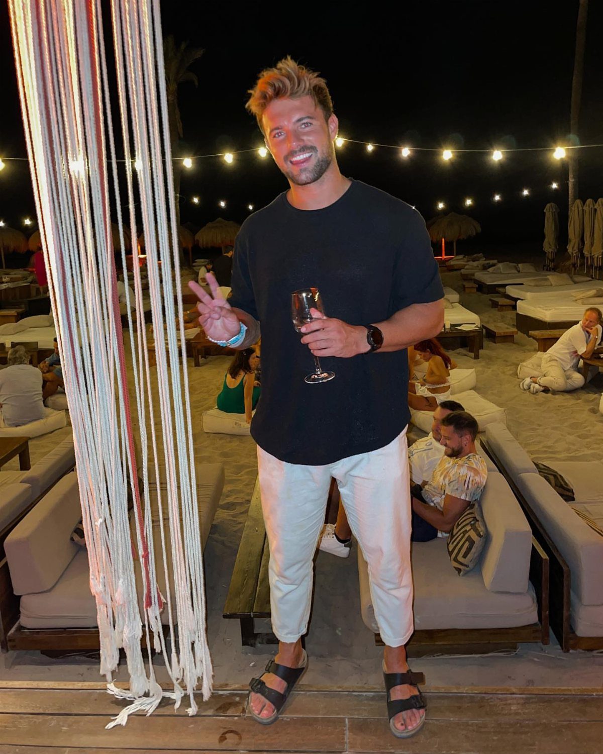 Smiling man in black t-shirt and white trousers at a beach lounge.