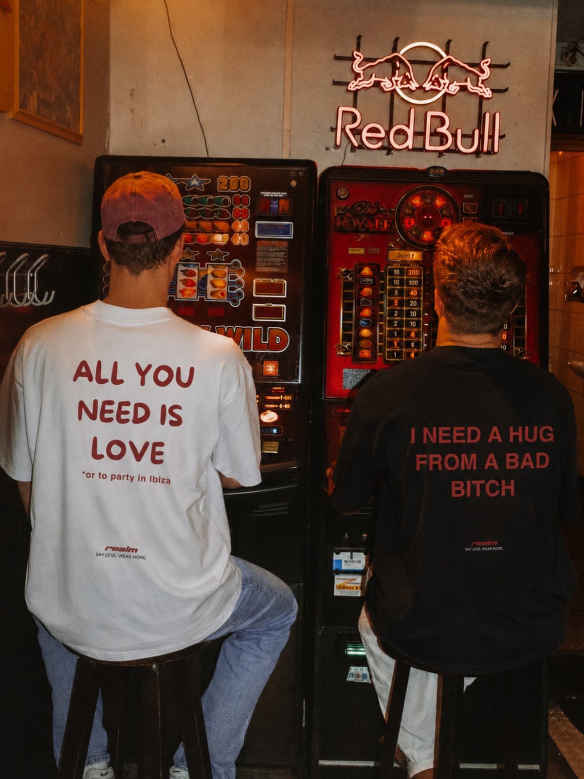Two men wearing bold graphic t-shirts sitting at slot machines in a bar.