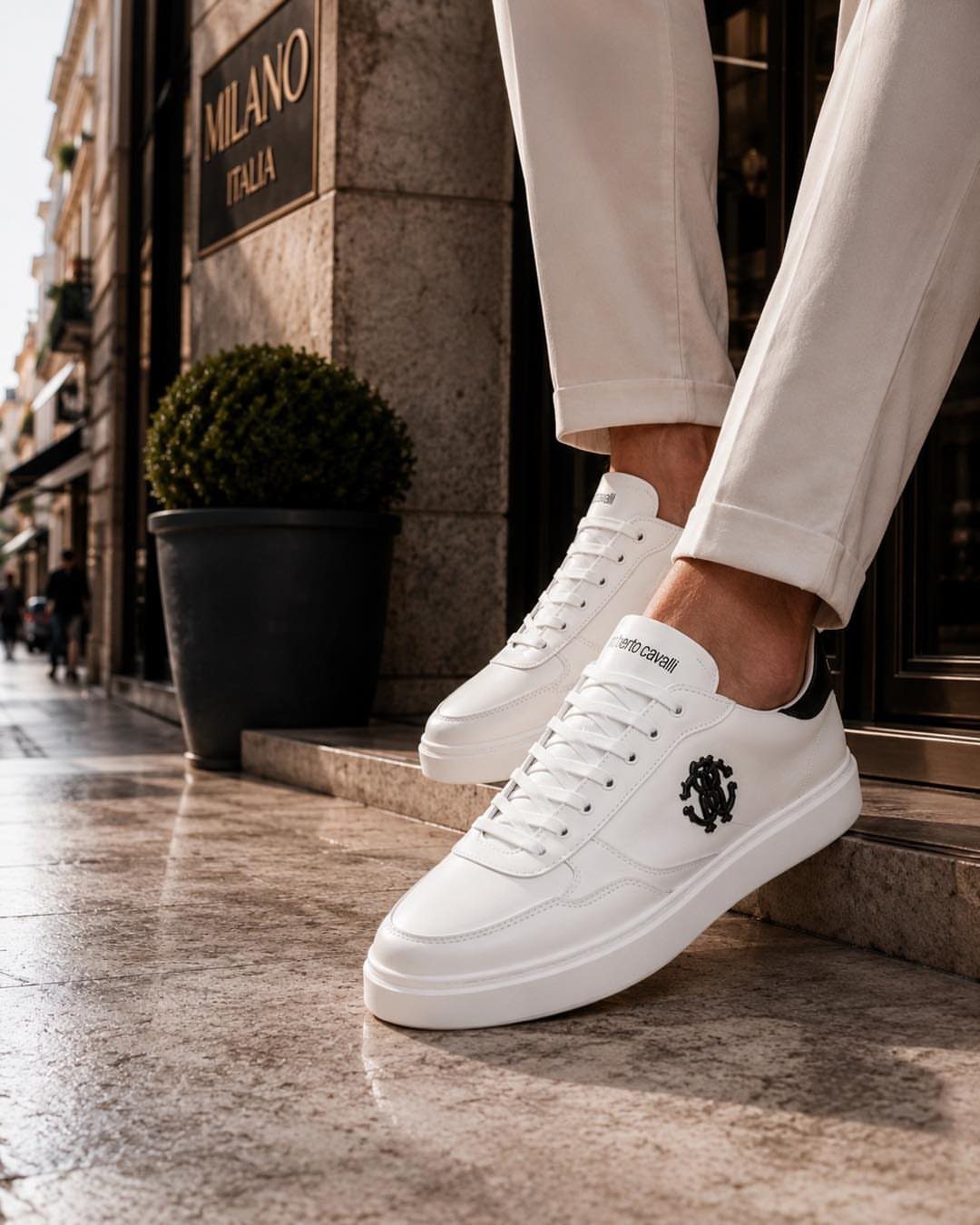 White leather sneakers with black logo and white trousers on stone pavement.