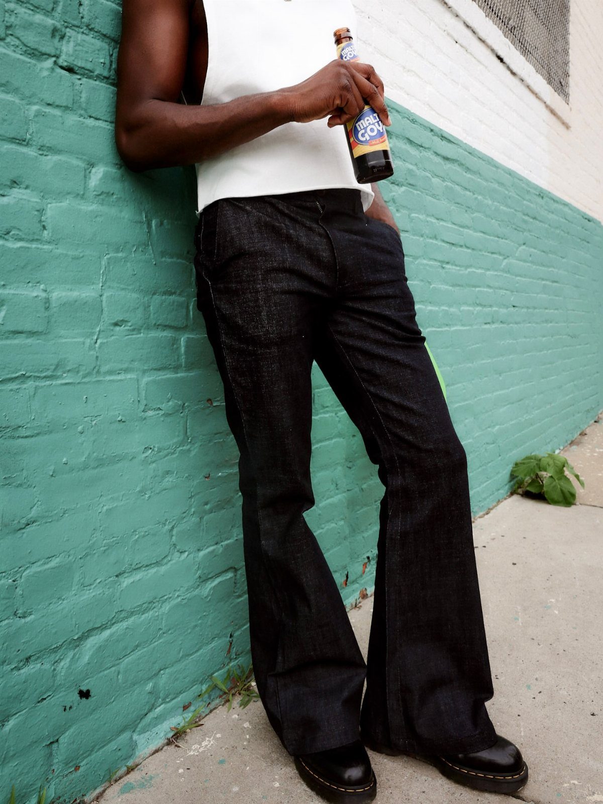 10 Images of 70's Men's Fashion from Flares to Fur Coats 2 Person wearing dark denim bell-bottom pants and a white top leaning against teal wall.
