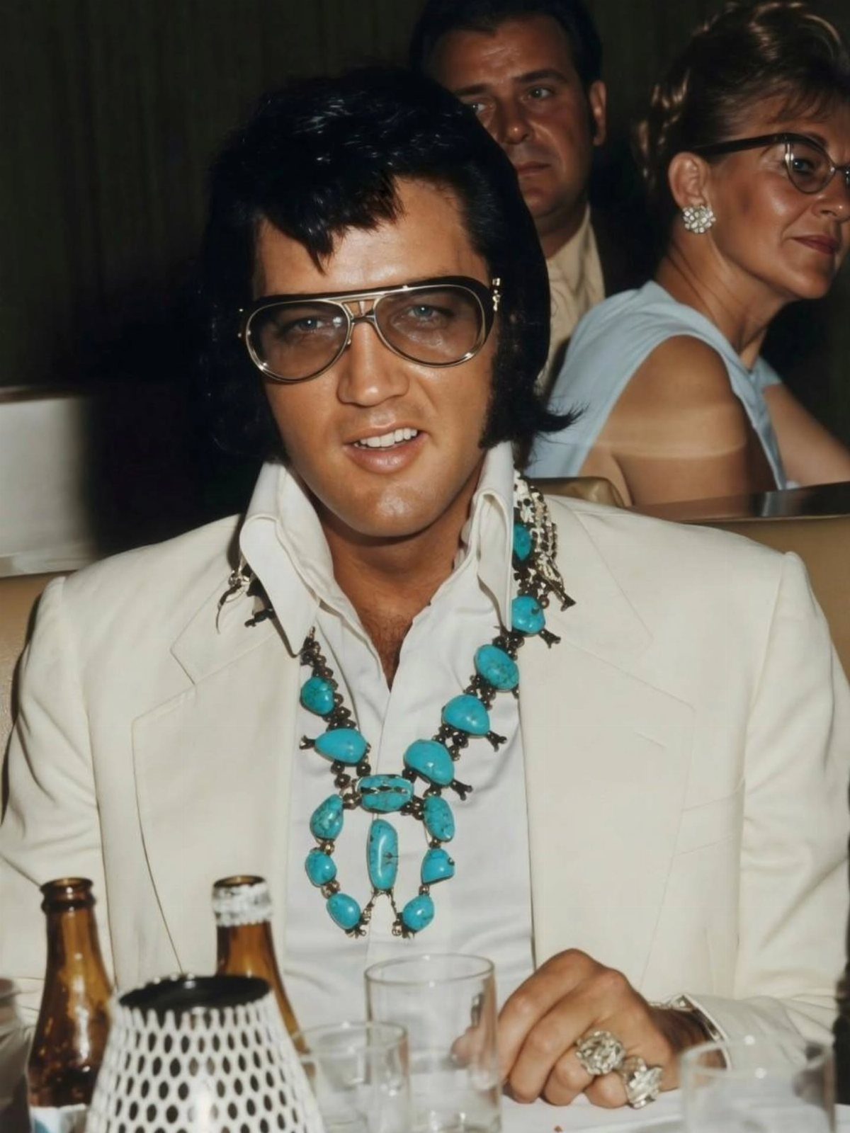 10 Images of 70's Men's Fashion from Flares to Fur Coats 5 Man wearing oversized gold-rimmed sunglasses, white suit, and large turquoise necklace.