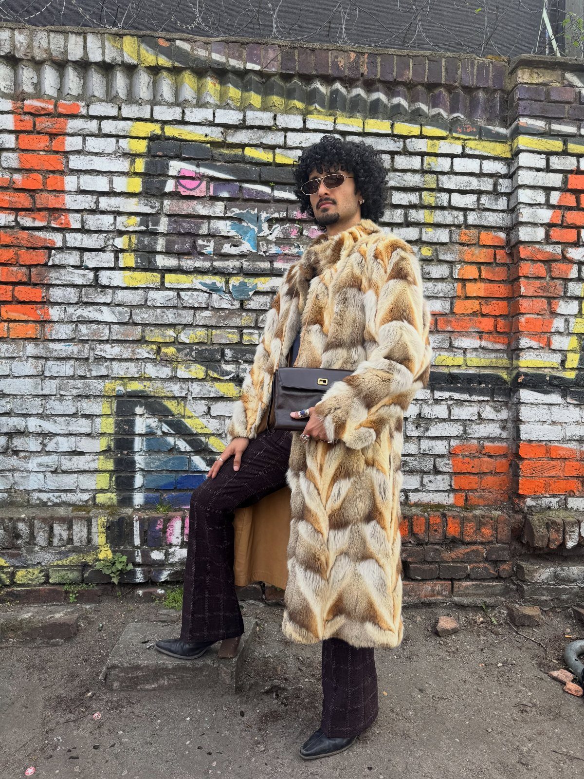 10 Images of 70's Men's Fashion from Flares to Fur Coats 6 Man in a long faux-fur coat and flared trousers posing by a graffiti wall.
