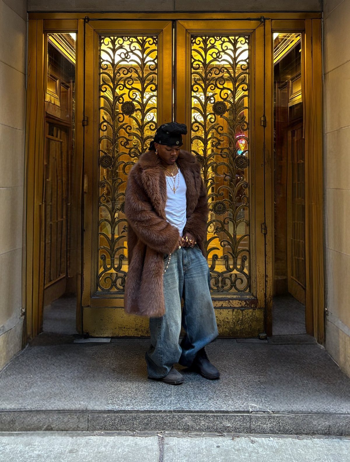 Man in brown fur coat and baggy jeans posing before gold doors.
