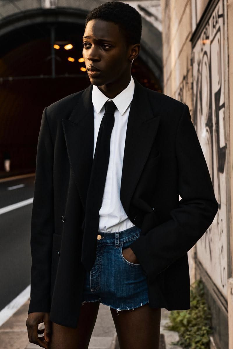 Man in oversized black blazer, white shirt, black tie, and low-rise denim mini shorts.