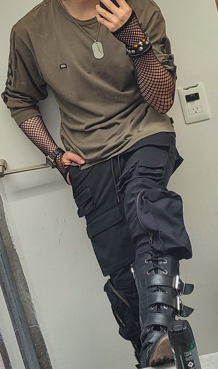 Person in olive shirt, fishnet sleeves, black cargo pants, and heavy buckled boots.