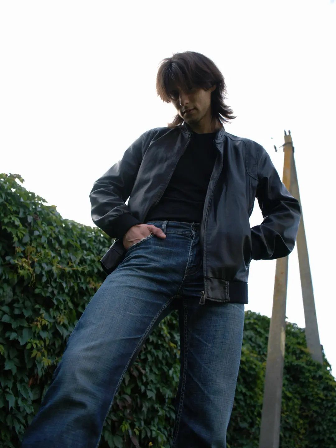Man wearing a dark bomber jacket and blue jeans standing by a hedge.
