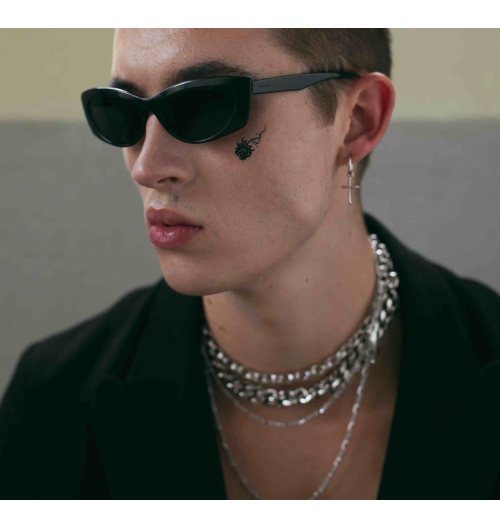 Man wearing dark tinted sunglasses and layered silver chain necklaces with a black blazer.