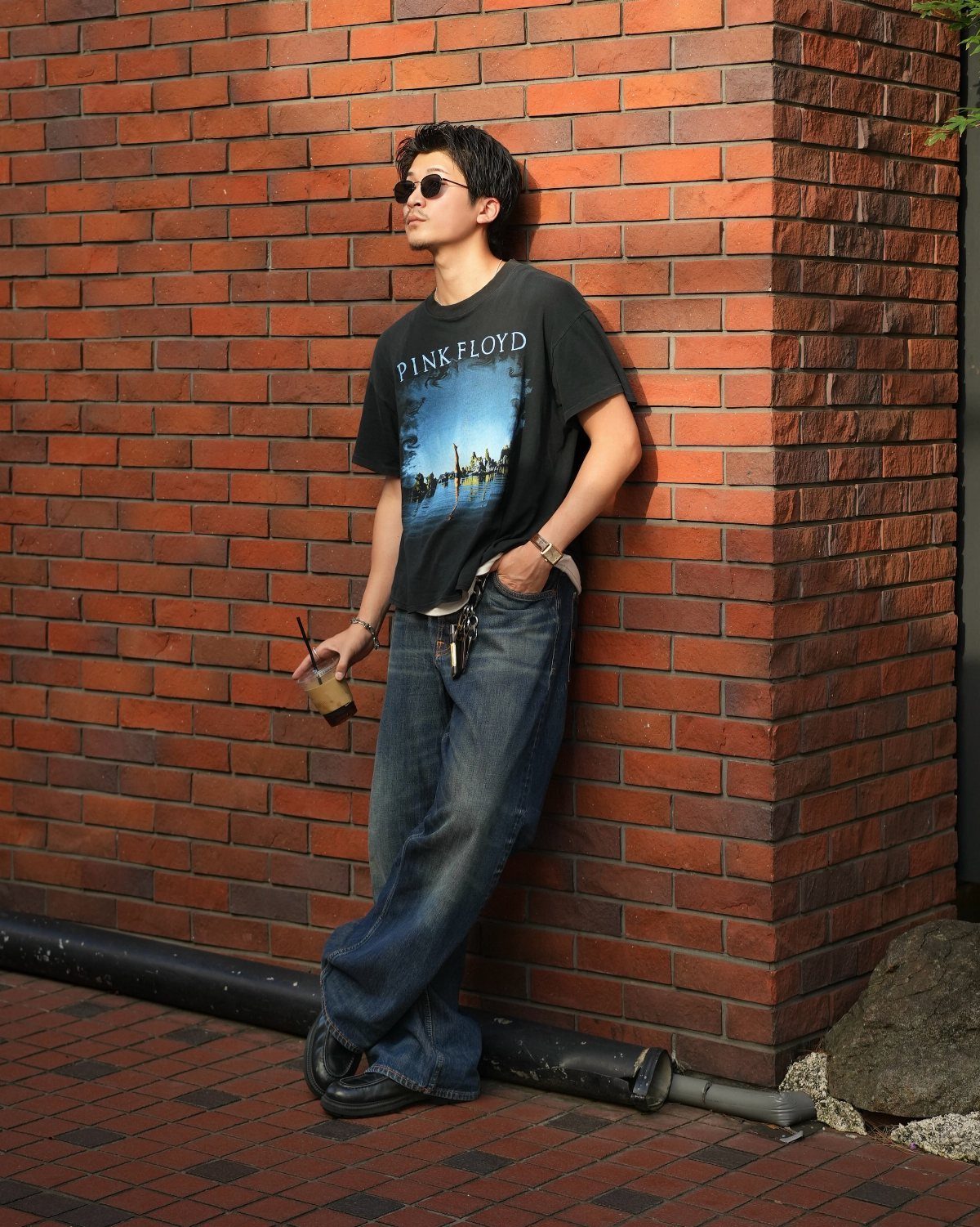 Man in Pink Floyd band tee and baggy jeans leaning against brick wall.