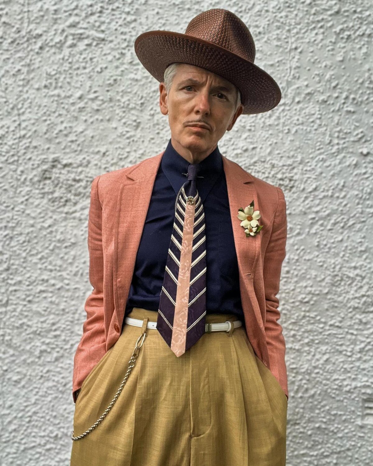 Man in a salmon blazer, tan high-waisted trousers, and brown wide-brimmed hat.