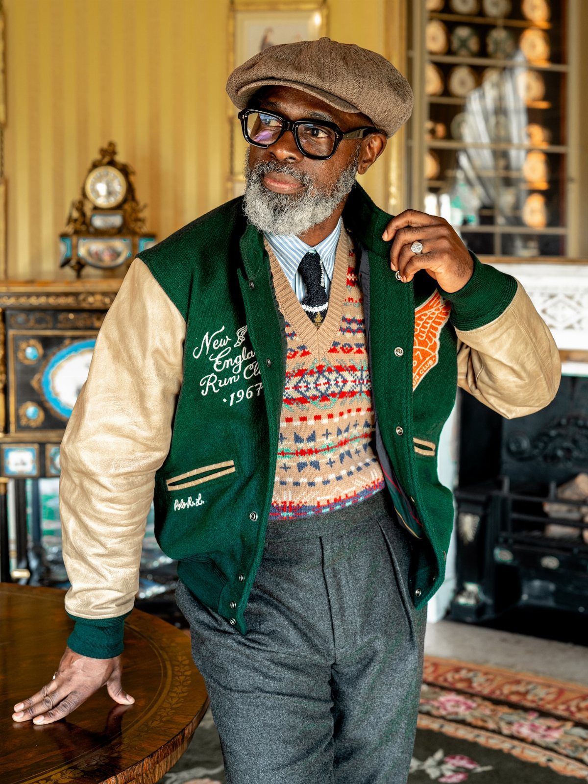 Man in vintage attire wearing a varsity jacket, patterned sweater, and brown flat cap.