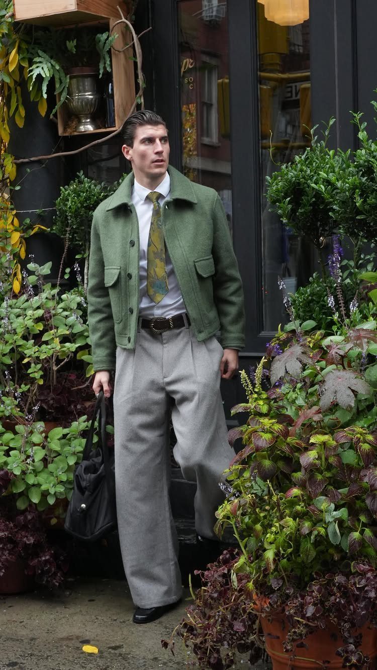 Man in green jacket and wide grey trousers posing among lush city plants.
