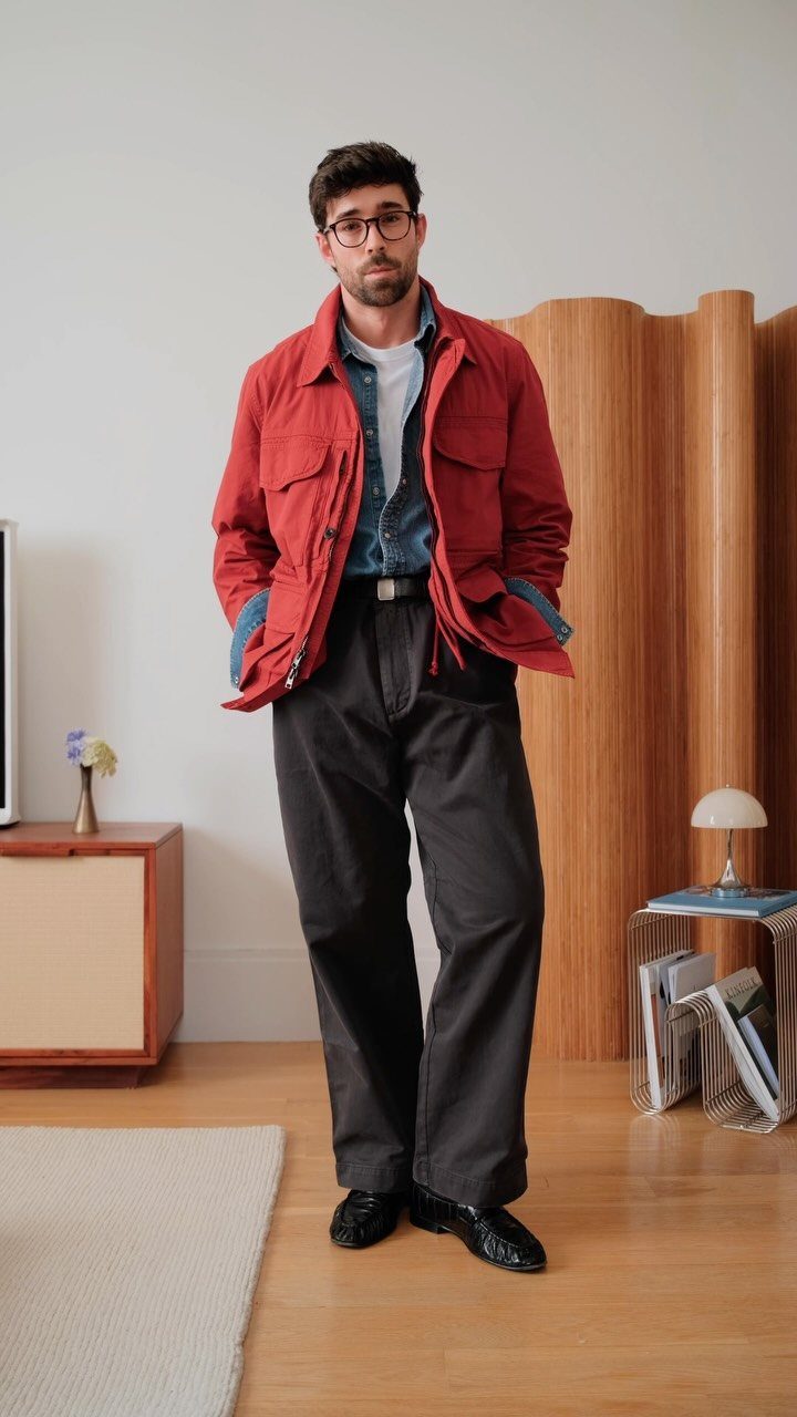 Man wearing a red utility jacket, blue shirt, and dark trousers in a modern room.