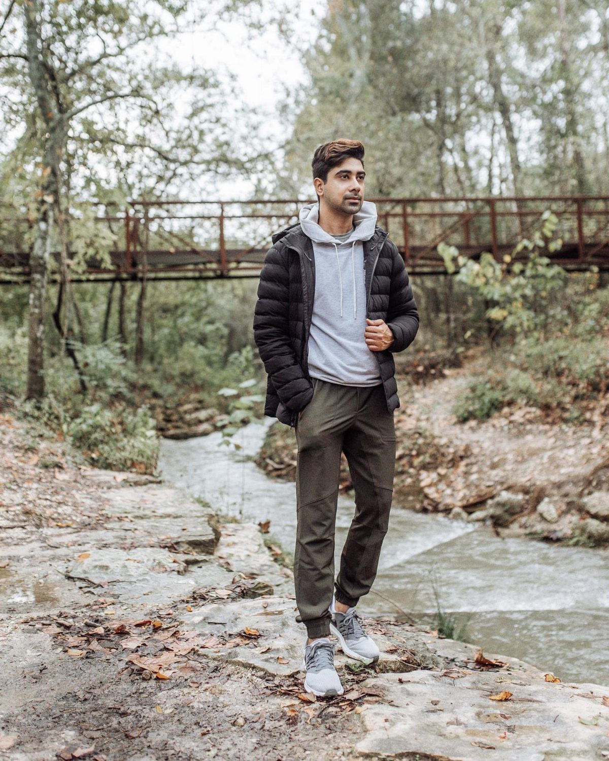 Man in black puffer jacket and olive joggers standing by a forest stream.
