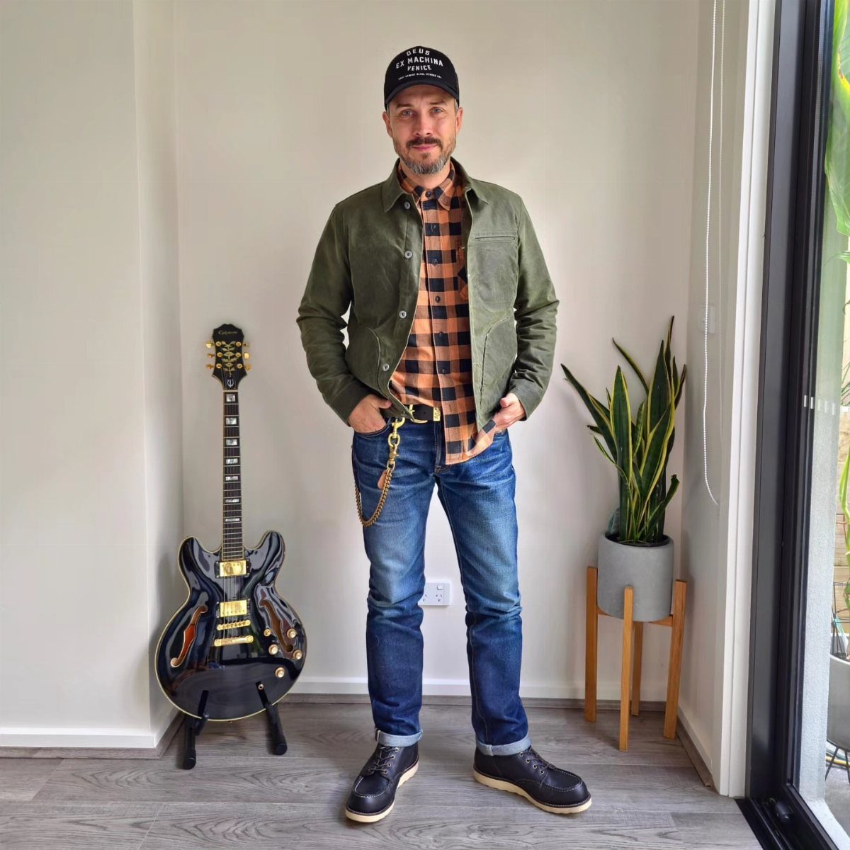 Man wearing olive jacket, plaid shirt, and blue jeans standing next to electric guitar.