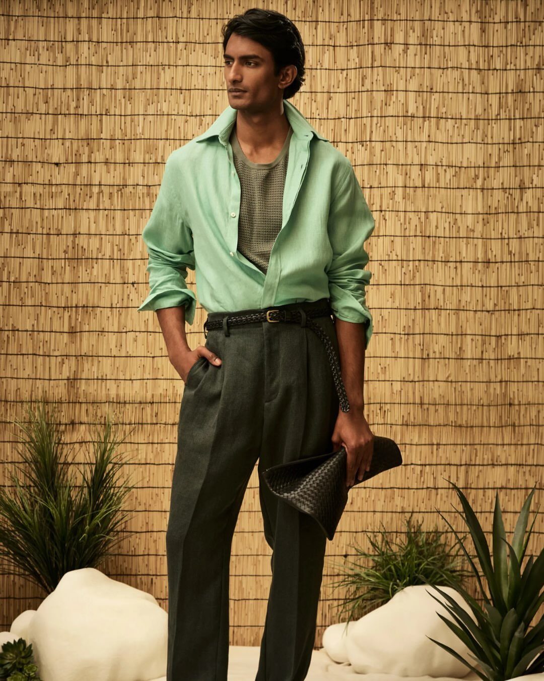Man in a light green shirt and high-waisted grey trousers posing against a woven wall.