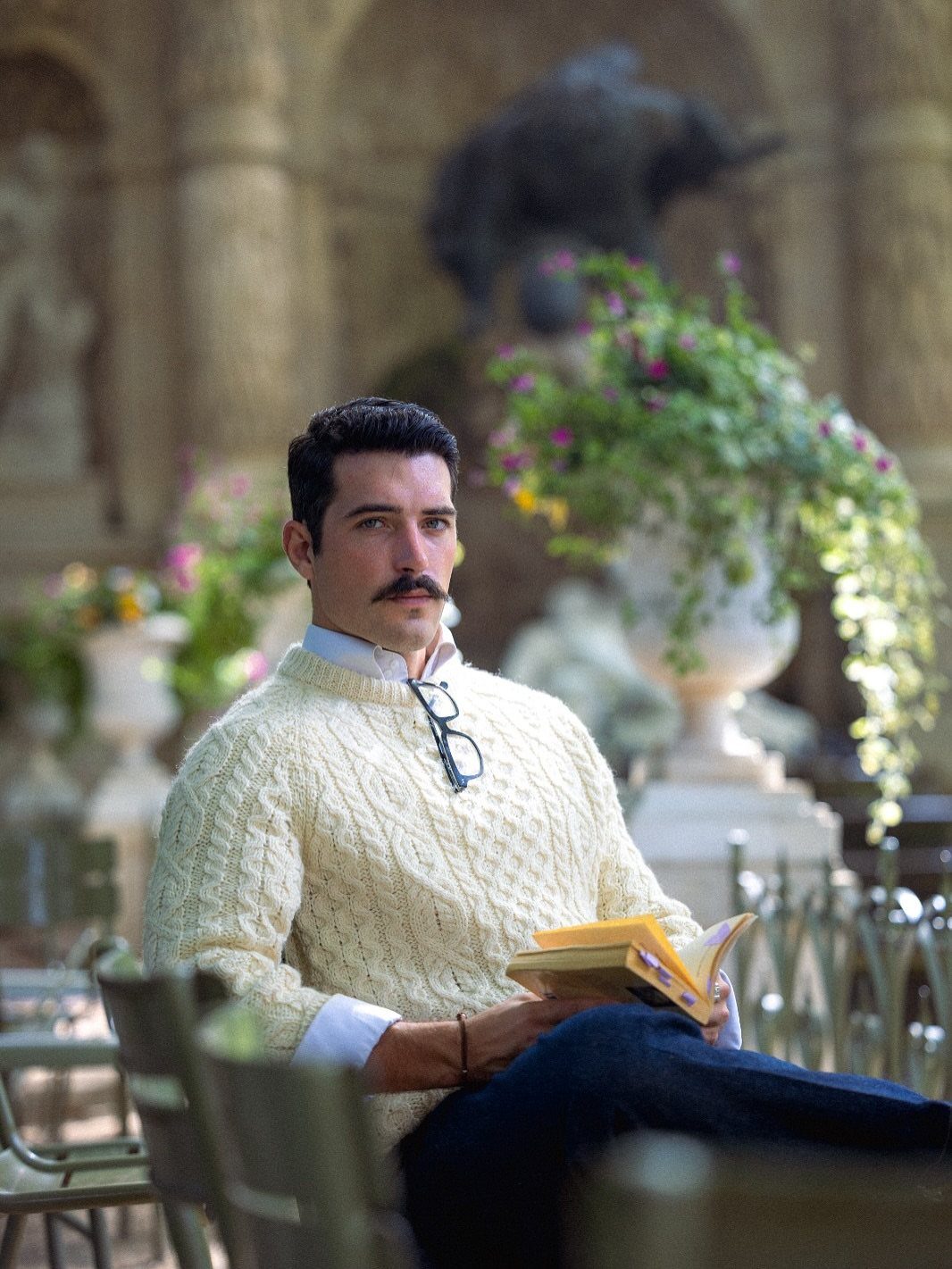 Man in cream cable-knit sweater and dark trousers reading a book in a garden.