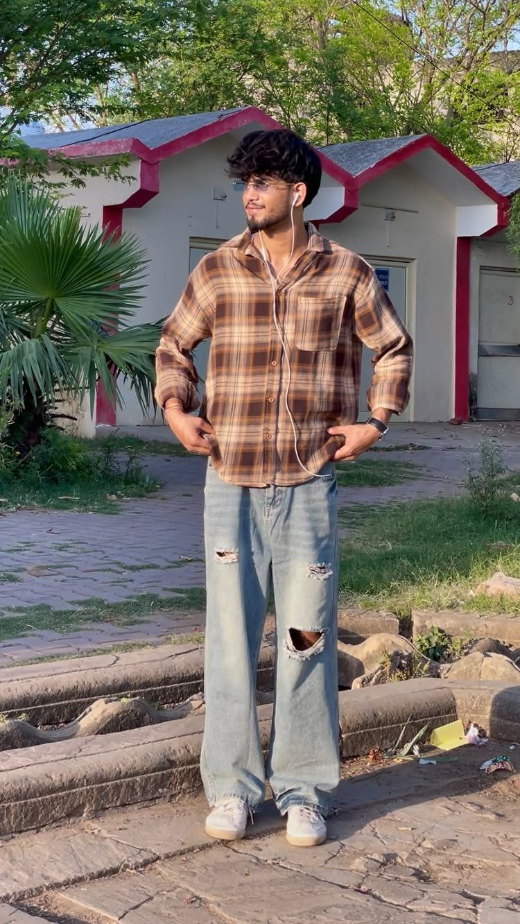 Man wearing a brown checkered shirt and ripped light blue jeans standing outdoors.