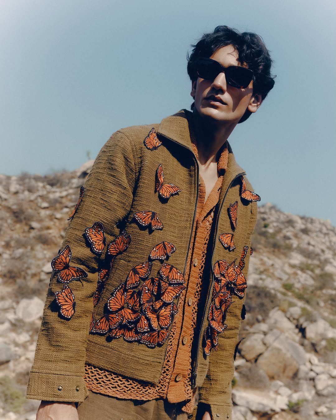 Man in a brown jacket covered in orange butterfly patches standing in a rocky area.