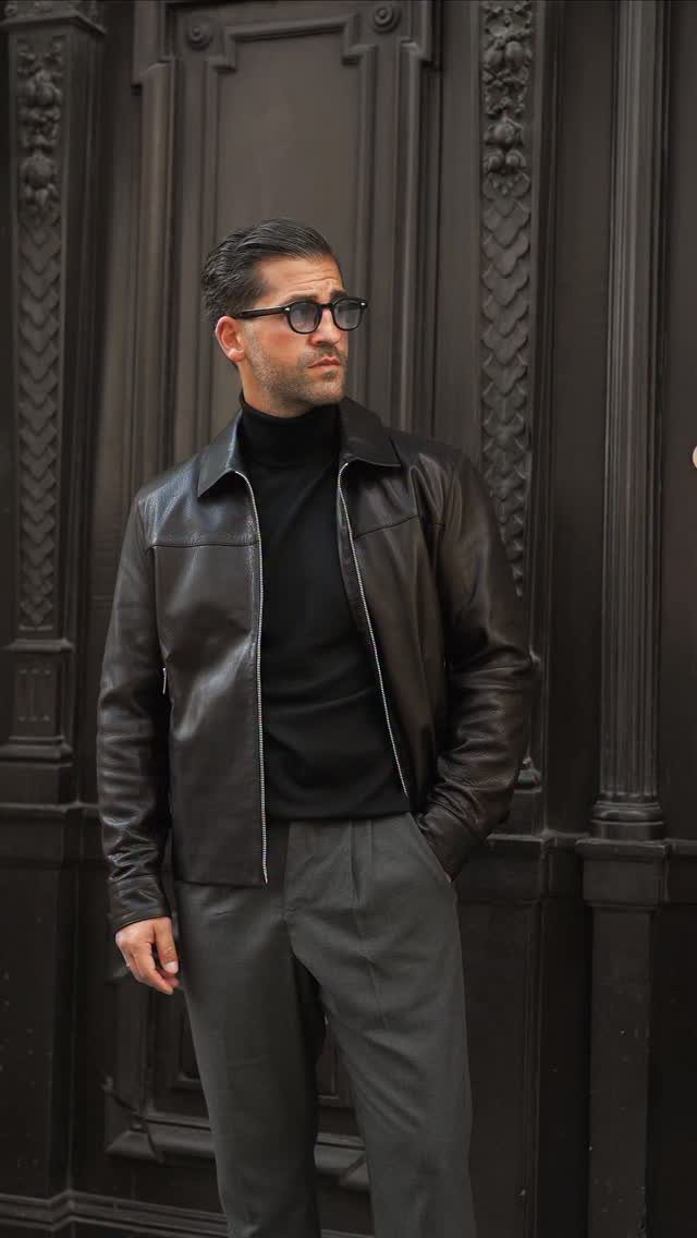 Man wearing a black turtleneck under a brown leather jacket and grey trousers.