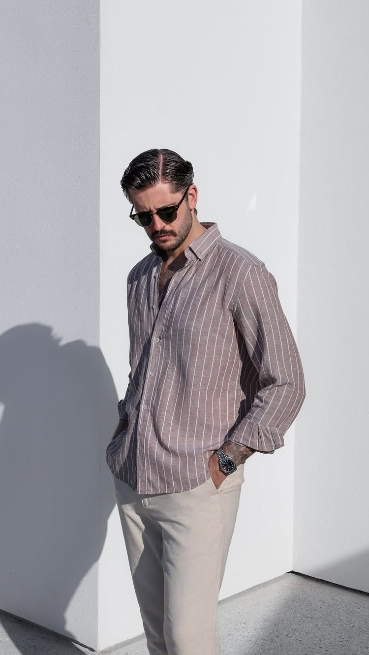 Man in striped brown shirt and beige trousers posing against a white wall.