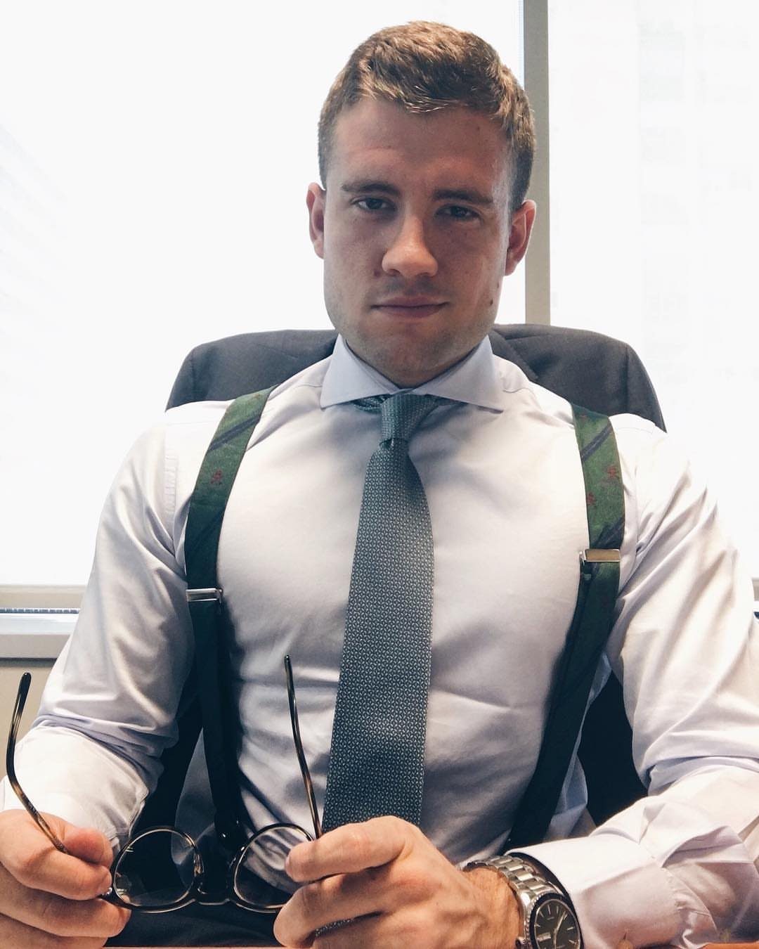 Man in white dress shirt, grey tie, and green suspenders posing in office.