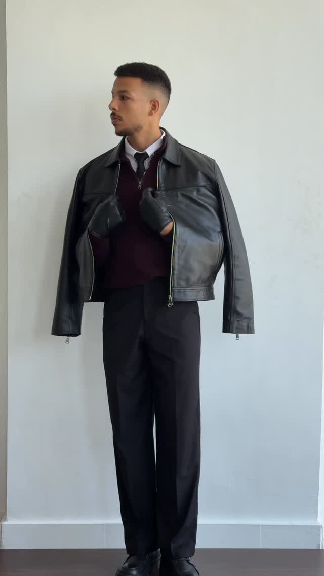 Man wearing a leather jacket, burgundy sweater, and black trousers against a white wall.