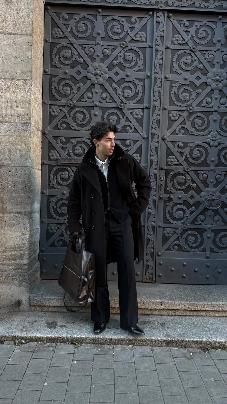 Man in black overcoat and trousers holding a leather bag by a metal door