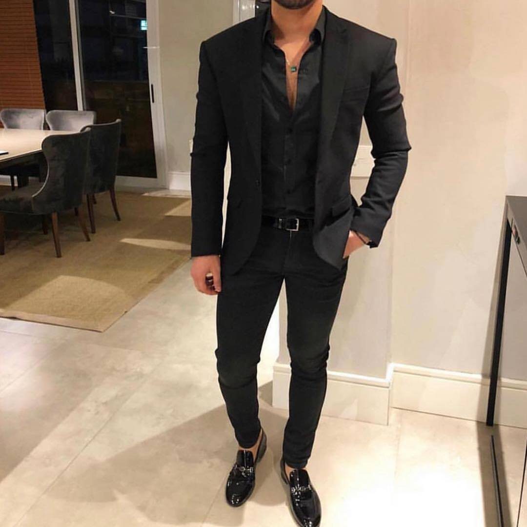Man wearing a monochrome all-black suit with a blazer and loafers in a room.