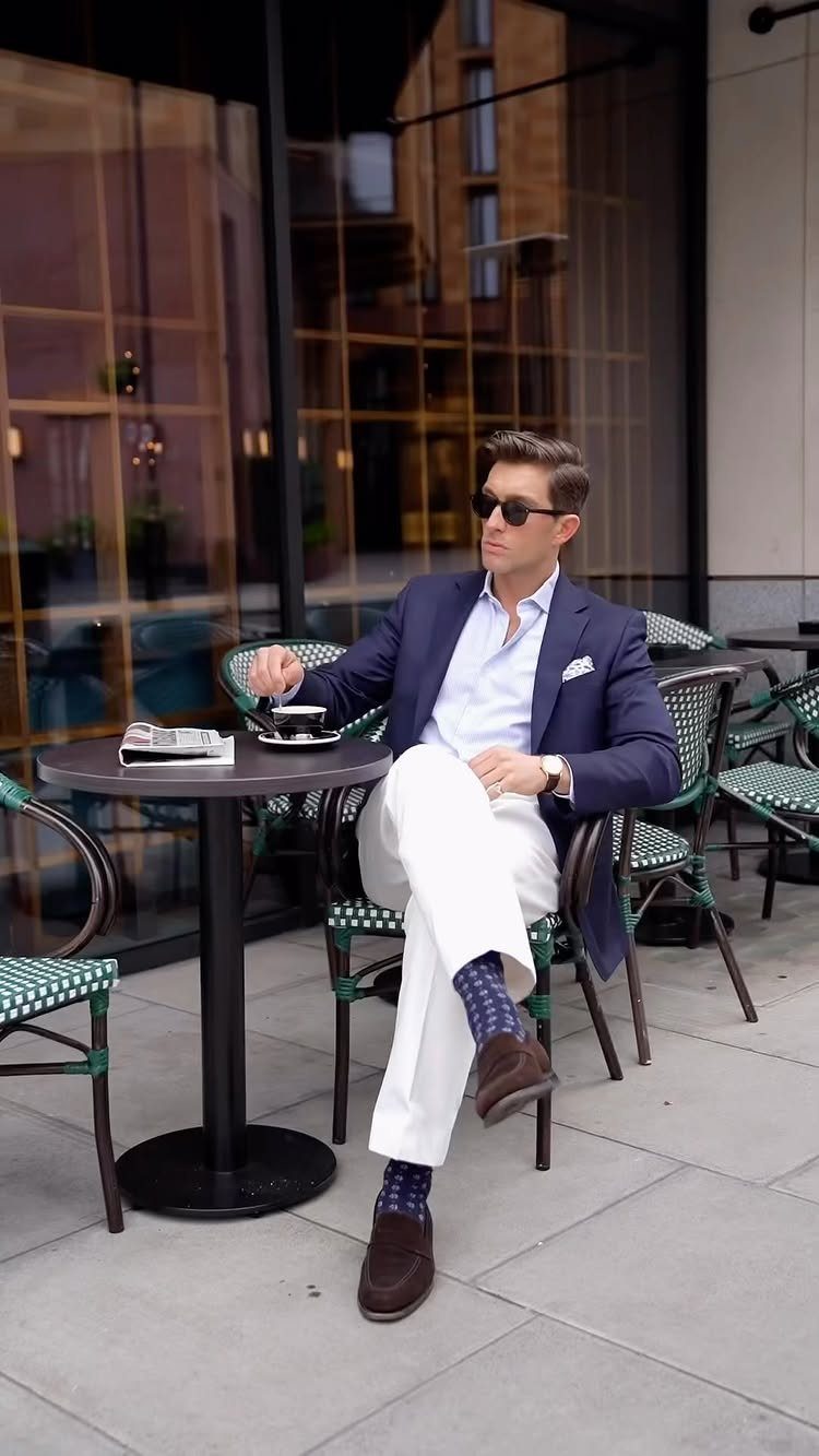 Man in navy blazer and white trousers sitting at an outdoor cafe table.