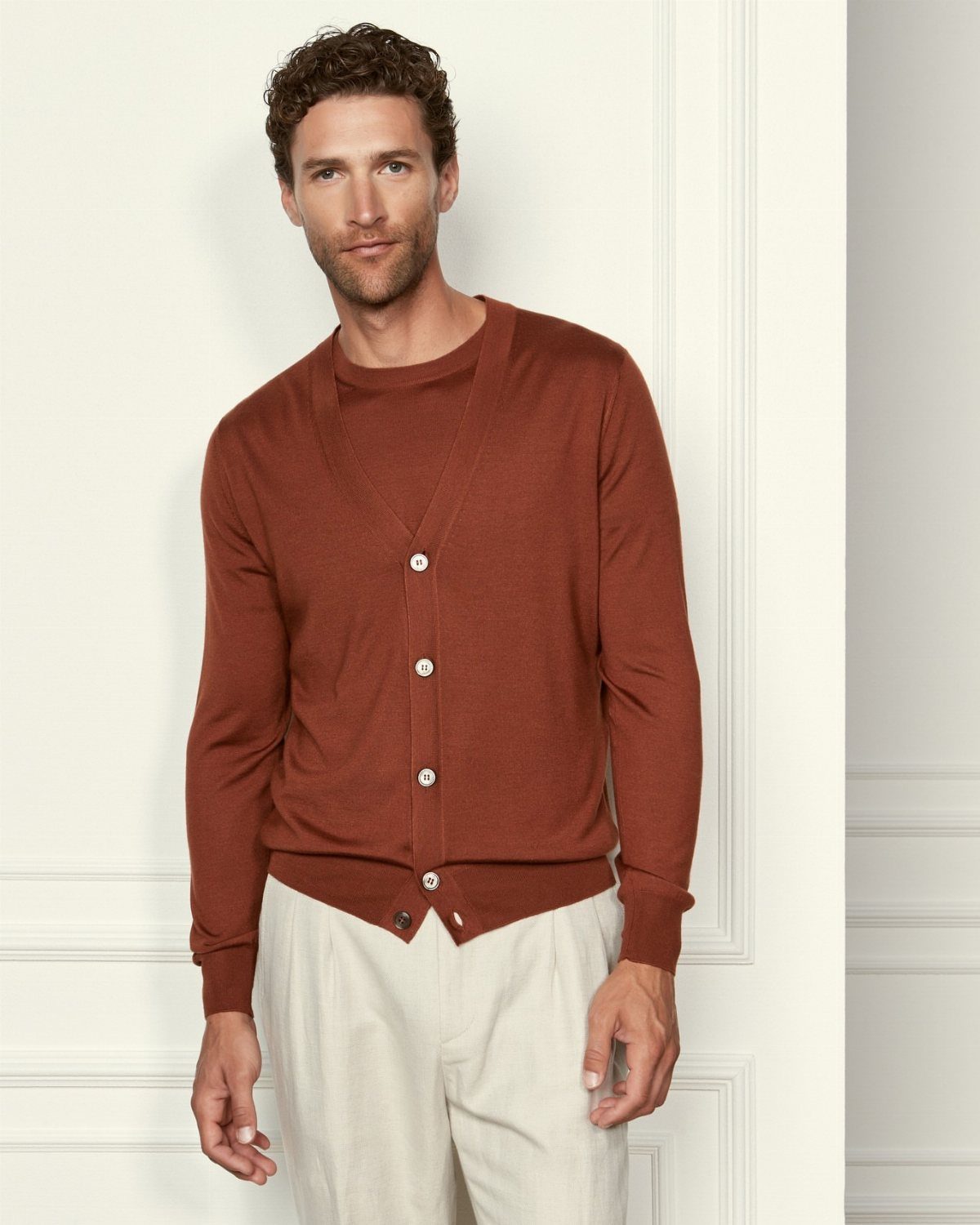Man wearing a rust-colored knit cardigan and off-white trousers against a white wall.