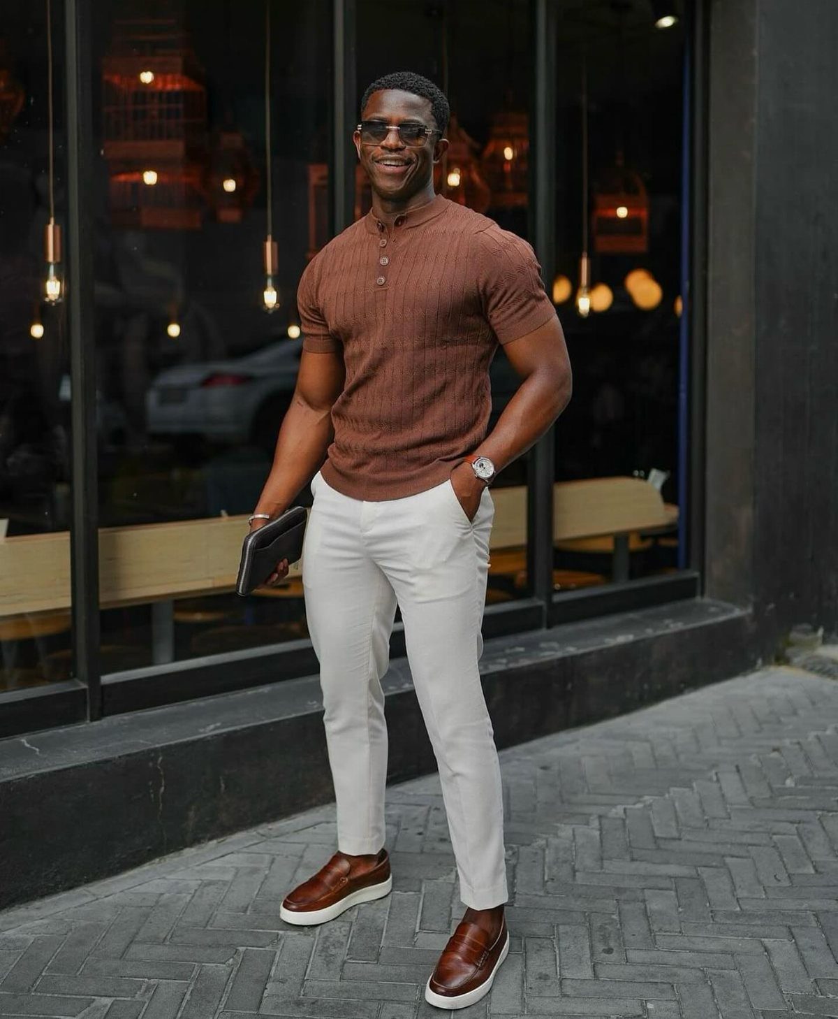Man wearing a brown ribbed shirt, white trousers, and brown shoes standing outdoors.
