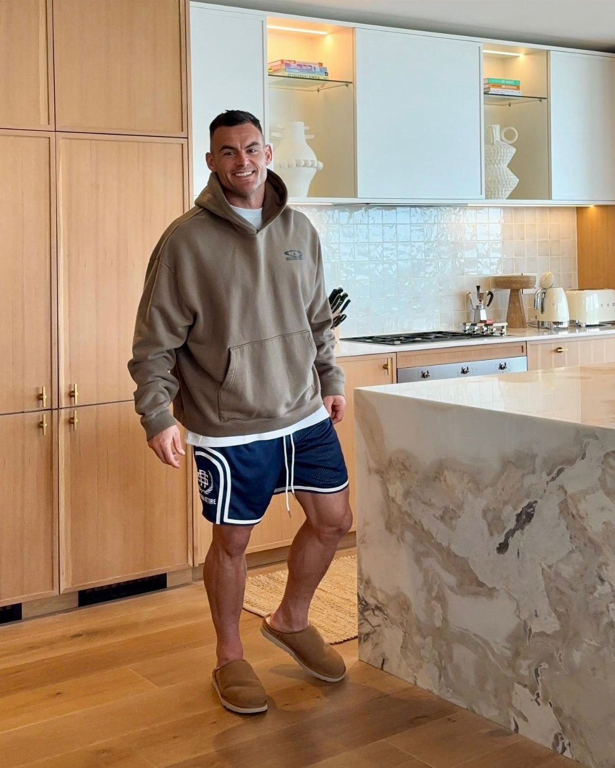 Man wearing tan hoodie, navy blue shorts, and tan suede clogs in a kitchen.