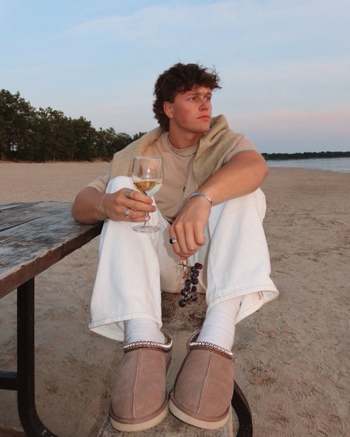 Man in white pants and tan Ugg Tasman slides sitting on a beach.