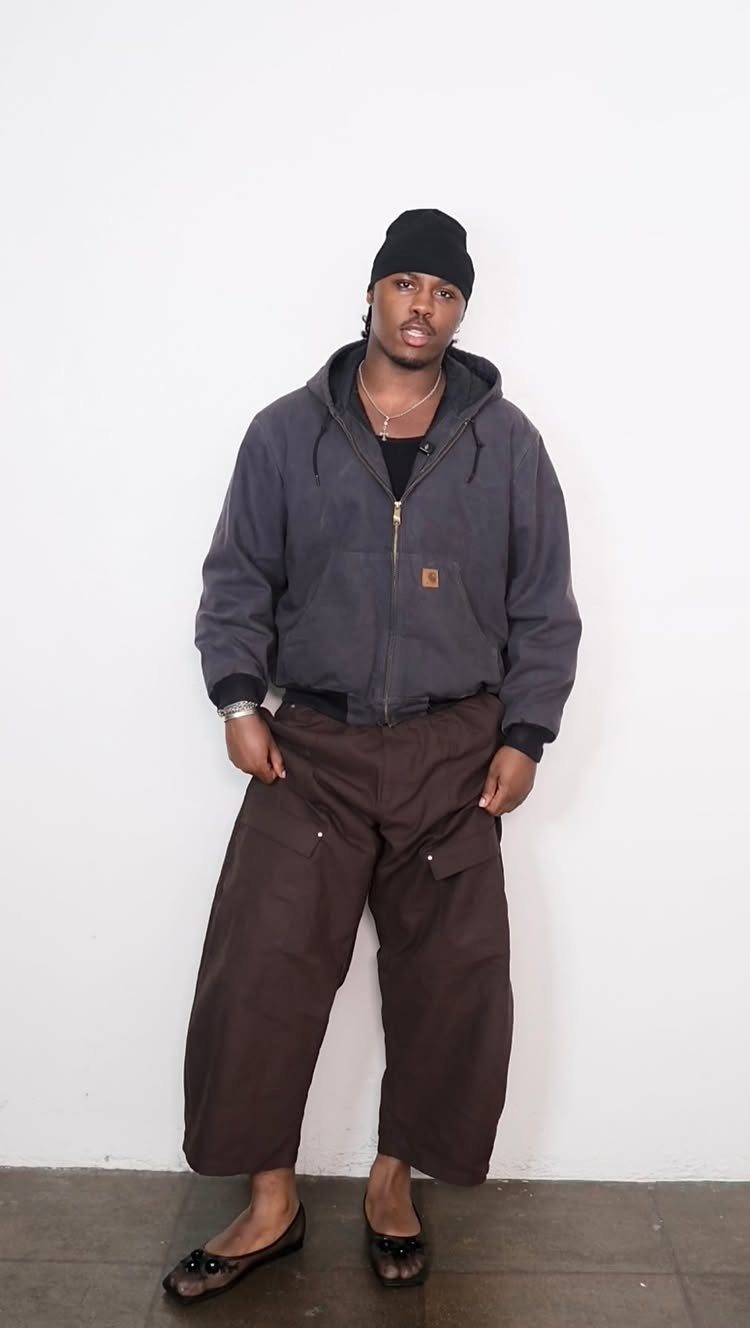 Man in oversized brown cargo pants and black ballet flats posing against white wall